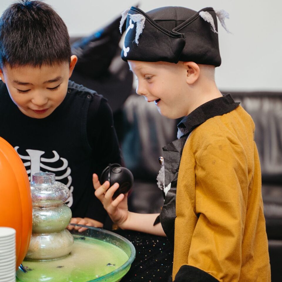 Two kids dressed in Halloween costumes excitedly explore a bubbling green potion with spooky props like a pirate hat and skeleton outfit, surrounded by magical Halloween vibes.