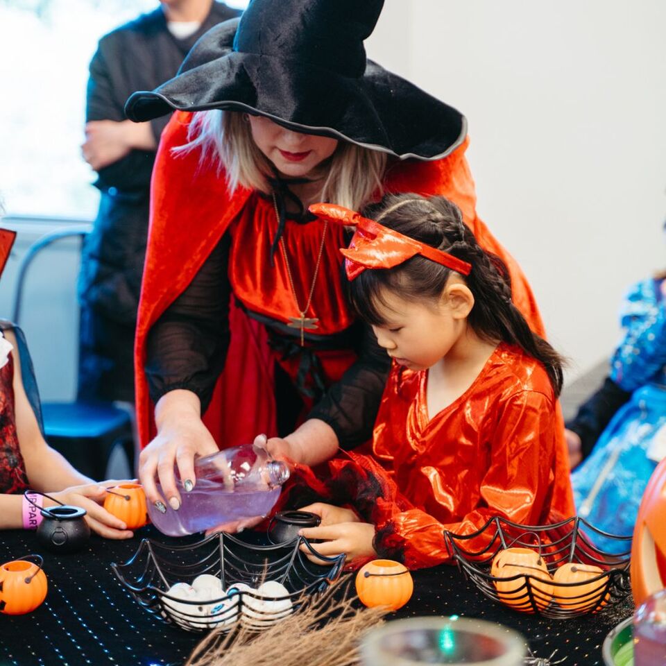 A magical moment as Glenda Goodspell, the Good Witch from Sparkle & Spell Co., teaches a young witch in a red dress how to brew a sparkling potion, with Halloween props scattered across the table.